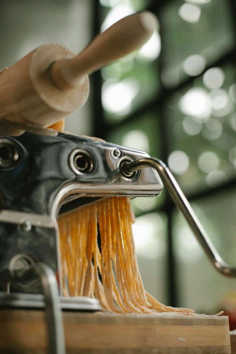 Metal modern pasta maker with raw cut noodles and rolling pin placed on wooden table in kitchen on blurred background
